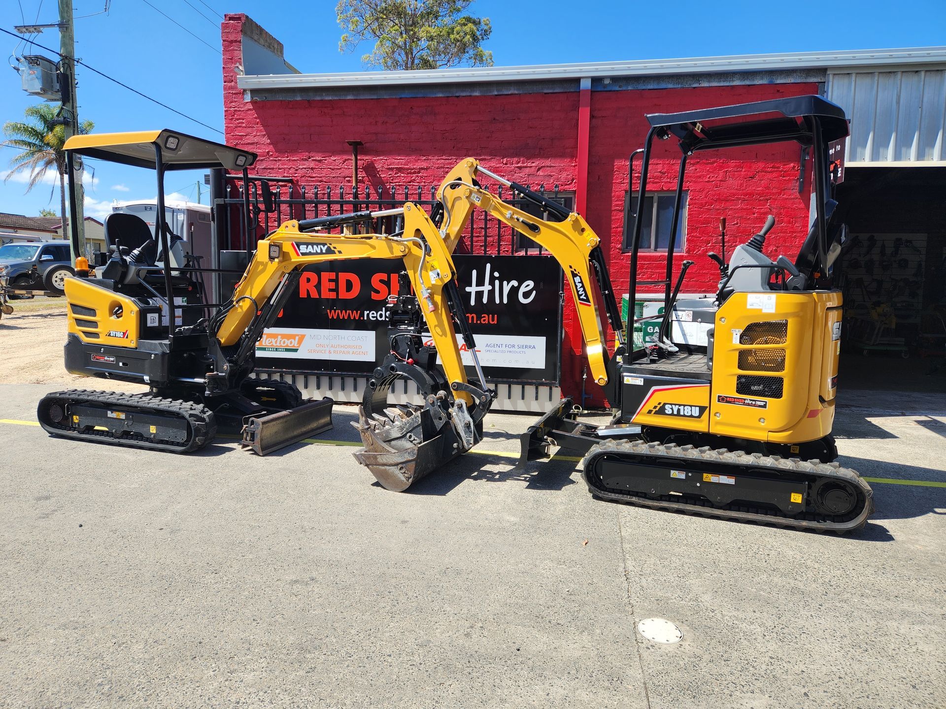 Two Small Excavators Are Parked In Front Of A Red Building — Red Shed Hire In Wauchope, NSW