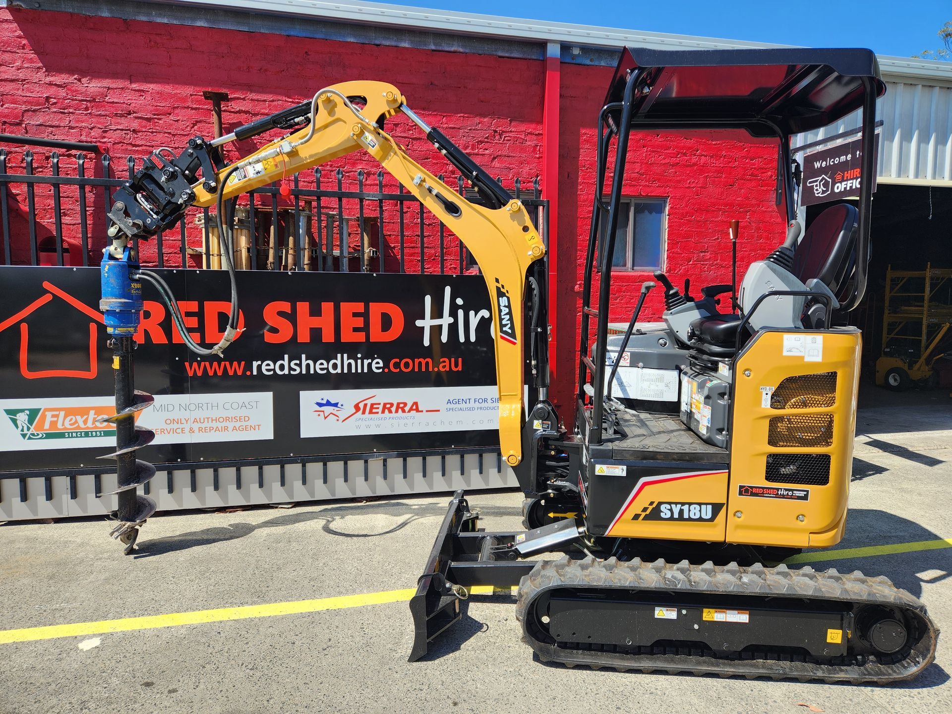 A Small Excavator Is Parked In Front Of A Red Shed Hire Sign — Red Shed Hire In Wauchope, NSW