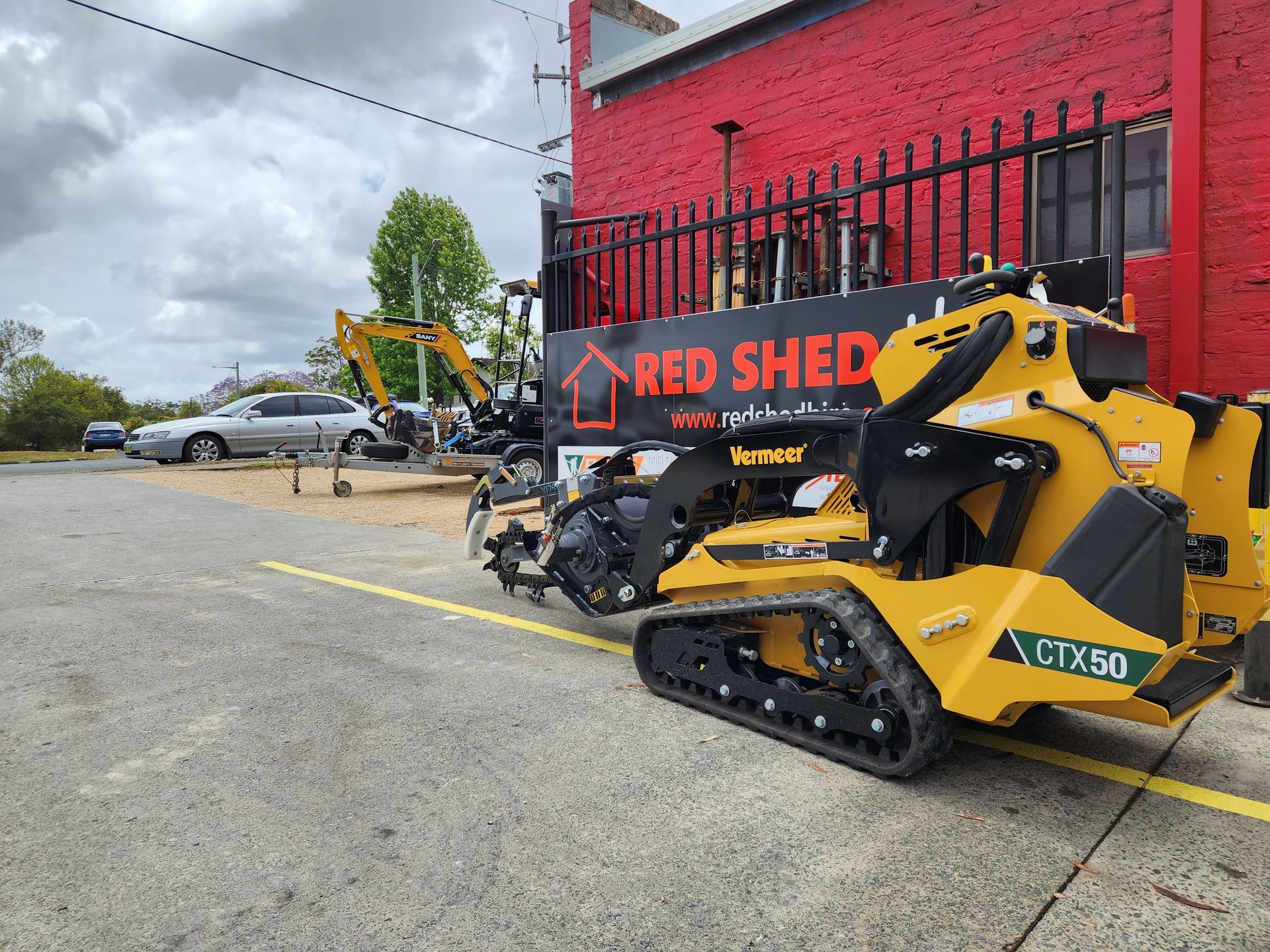A Yellow Tractor Is Parked In Front Of A Red Shed Building — Red Shed Hire In Wauchope, NSW