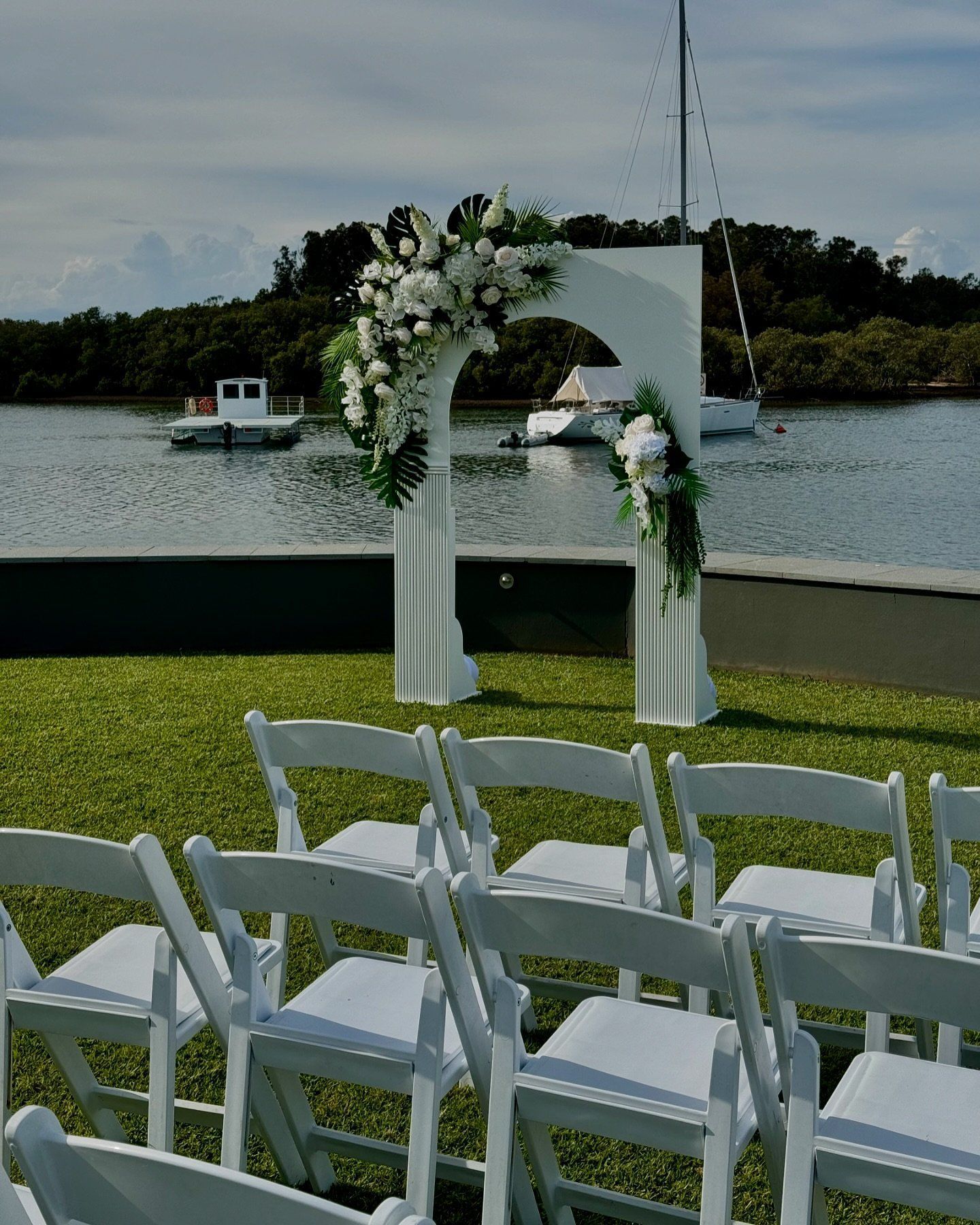 A Row Of White Folding Chairs Are Lined Up In Front Of A Floral Arch Overlooking A Body Of Water — Red Shed Hire In Wauchope, NSW