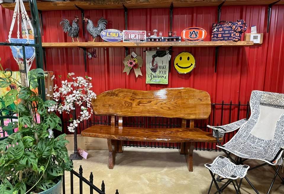 Wooden bench in front of a red wall with shelves holding decor. Plant, fence, and folding chair are visible.