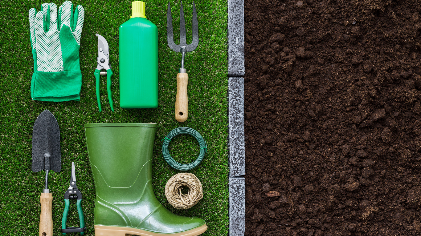 Gardening tools and boots arranged on grass next to a section of soil, ready for planting.