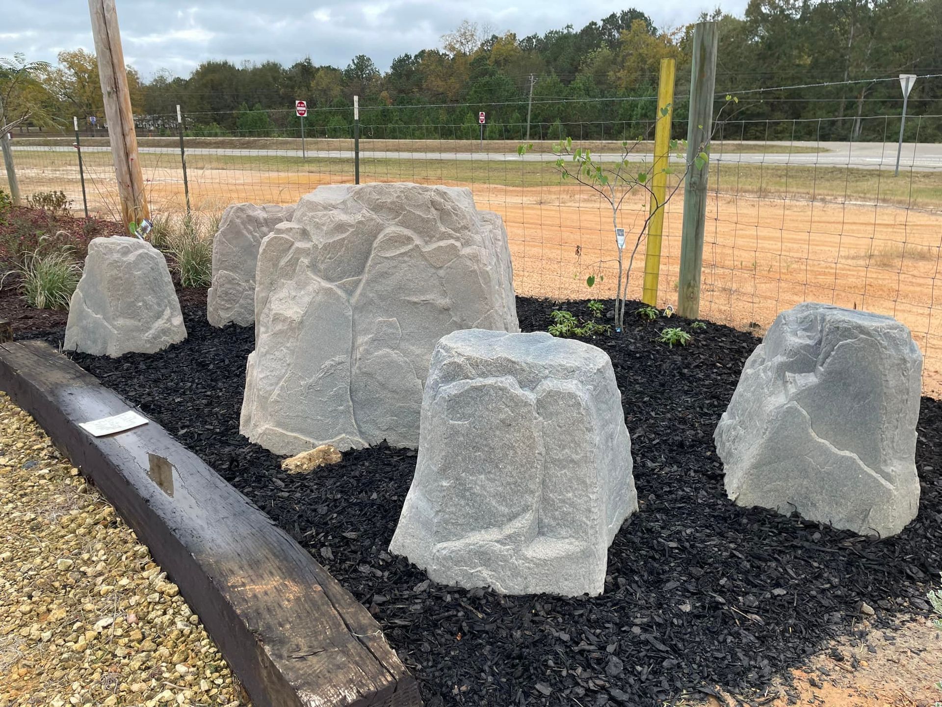 Gray rocks arranged in black mulch bed; wooden beam in foreground; background of trees and fence.