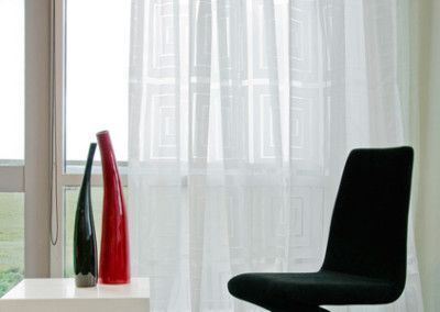 White sheer curtains frame a window with a table holding vases and a black chair.