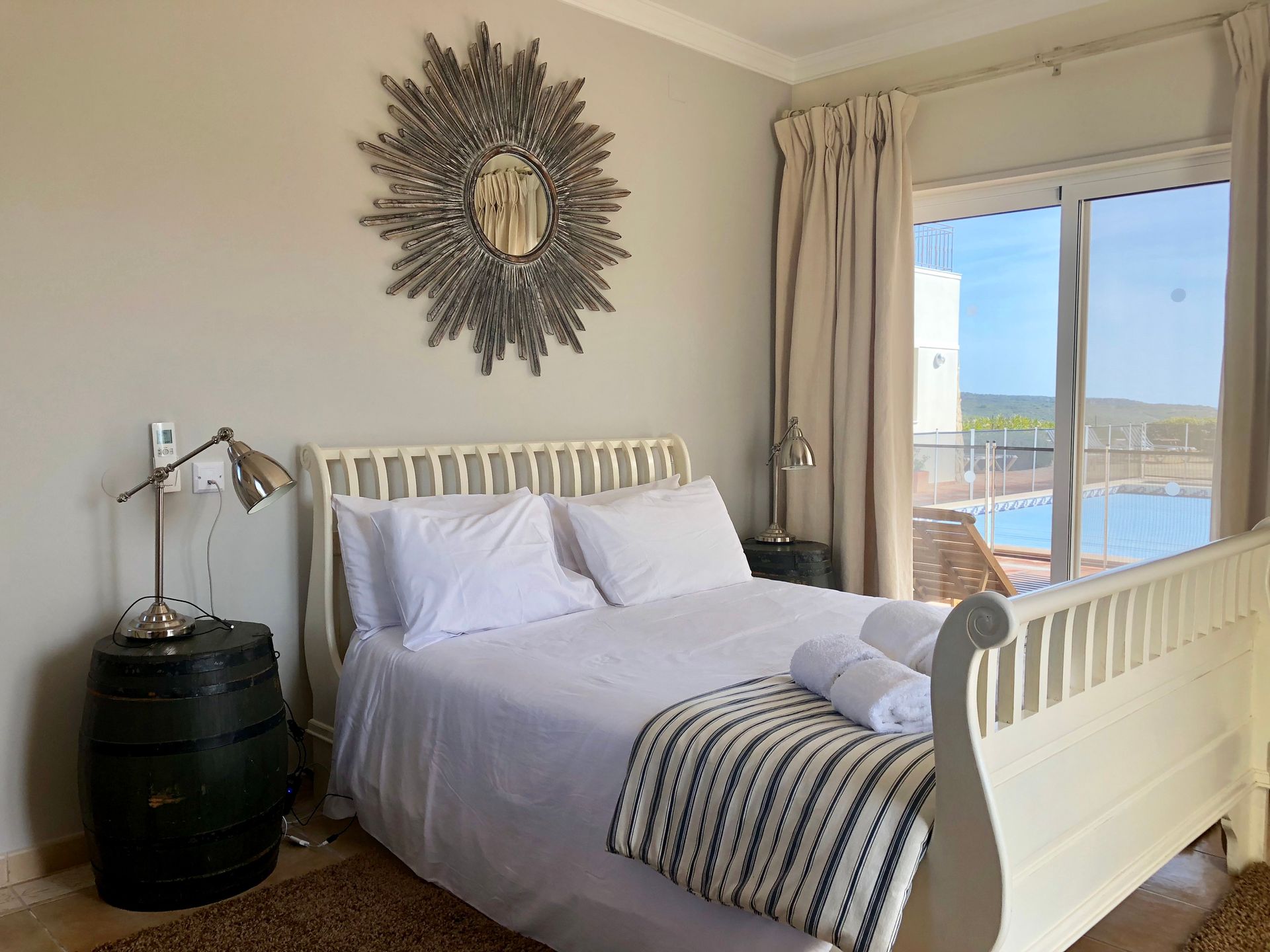 Bedroom with a white bed, gray wall, and large sunburst mirror. Sliding glass door with a pool view.
