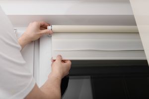 Person installing a white roller blind in a window frame. Person installing a cream-colored roller blind in a white window frame, with hands visible.