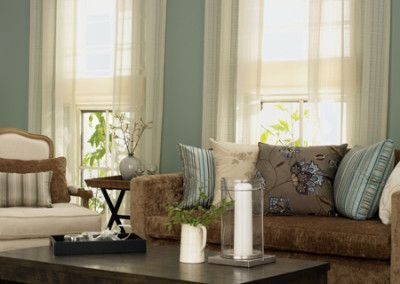 Living room with light blue walls, brown sofa and chair, two windows with sheer curtains.