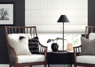 Two wooden armchairs with white cushions and pillows in a living room with a lamp and Roman shade.