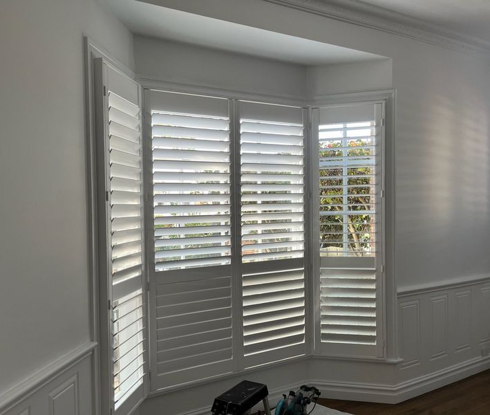 White window shutters in a bay window, partially open, with sunlight streaming through.