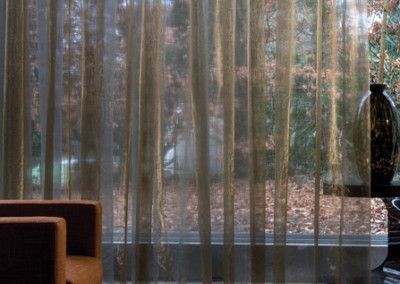 Sheer gold curtains in front of a window, with a dark vase and an orange chair visible.