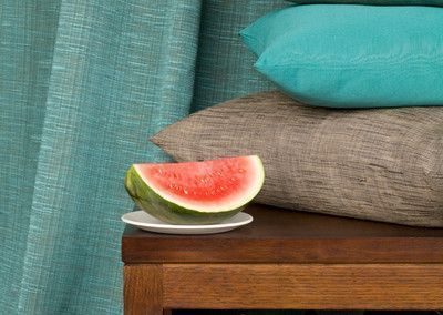 Watermelon slice on a small plate atop a wooden table, with pillows and blue curtains in the background.