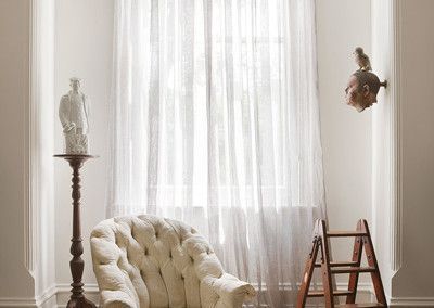 Cream-colored tufted chair, statue on stand, bust with bird, and ladder beside sheer curtained window in white room.