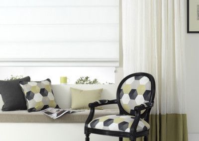 A sunny window seat with pillows and a patterned chair. White blinds, curtains, and a black accent.