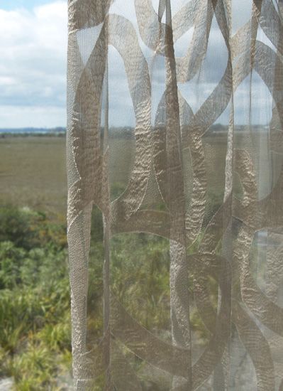 Sheer, tan curtain with an abstract design hangs in front of a green and brown landscape.