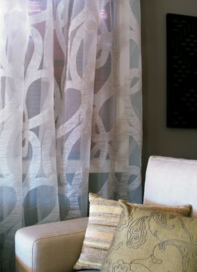 Sheer white curtains with a swirling pattern, beside a cream armchair with decorative pillows.
