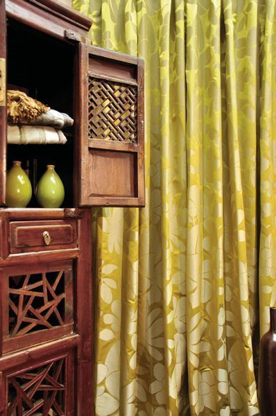 Wooden cabinet with open door revealing linens and vases, in front of yellow floral curtains.