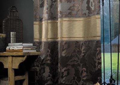 Dark floral curtain with a gold band, beside a rustic table and a view of a rainy landscape.