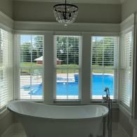 A white freestanding bathtub sits in front of a three-paneled window overlooking a backyard swimming pool.