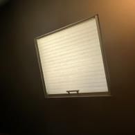 A square window with a closed, pleated white cellular shade set against a dark wall.
