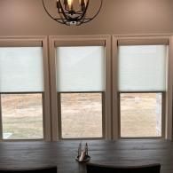 Three windows with light-filtering roller shades above a dark wooden table with a metal candle holder and a light fixture.