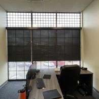 A modern office workstation with a wooden desk, black office chair, computer, and a grid-paned window with dark shades.