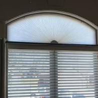 A white arched window shade with a fan pattern installed above horizontal white blinds.