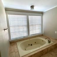 A beige tiled soaking tub sits below two windows with closed white blinds in a neutral-toned bathroom.