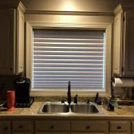 A kitchen sink with a dark faucet sits beneath a window covered by white pleated blinds, flanked by light-colored cabinets.