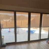Four large glass sliding doors covered by brown solar shades, showing a patio, yard, and fence outside.