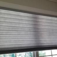A horizontal, pleated window shade in a light grey fabric, partially covering a window with visible outdoor greenery.