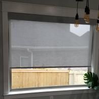 A white-framed window featuring a grey solar roller shade that covers the top half, revealing a view of a wooden fence.