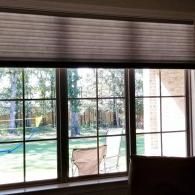 A large, multi-pane window featuring a partially closed, horizontal cellular shade, looking out onto a backyard.