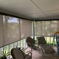 A screen-enclosed porch with light-filtering roller shades, two chairs, and a blue ladder in the foreground.