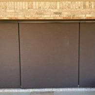 Dark brown solar shades are rolled down over glass panels beneath a light-colored brick exterior.
