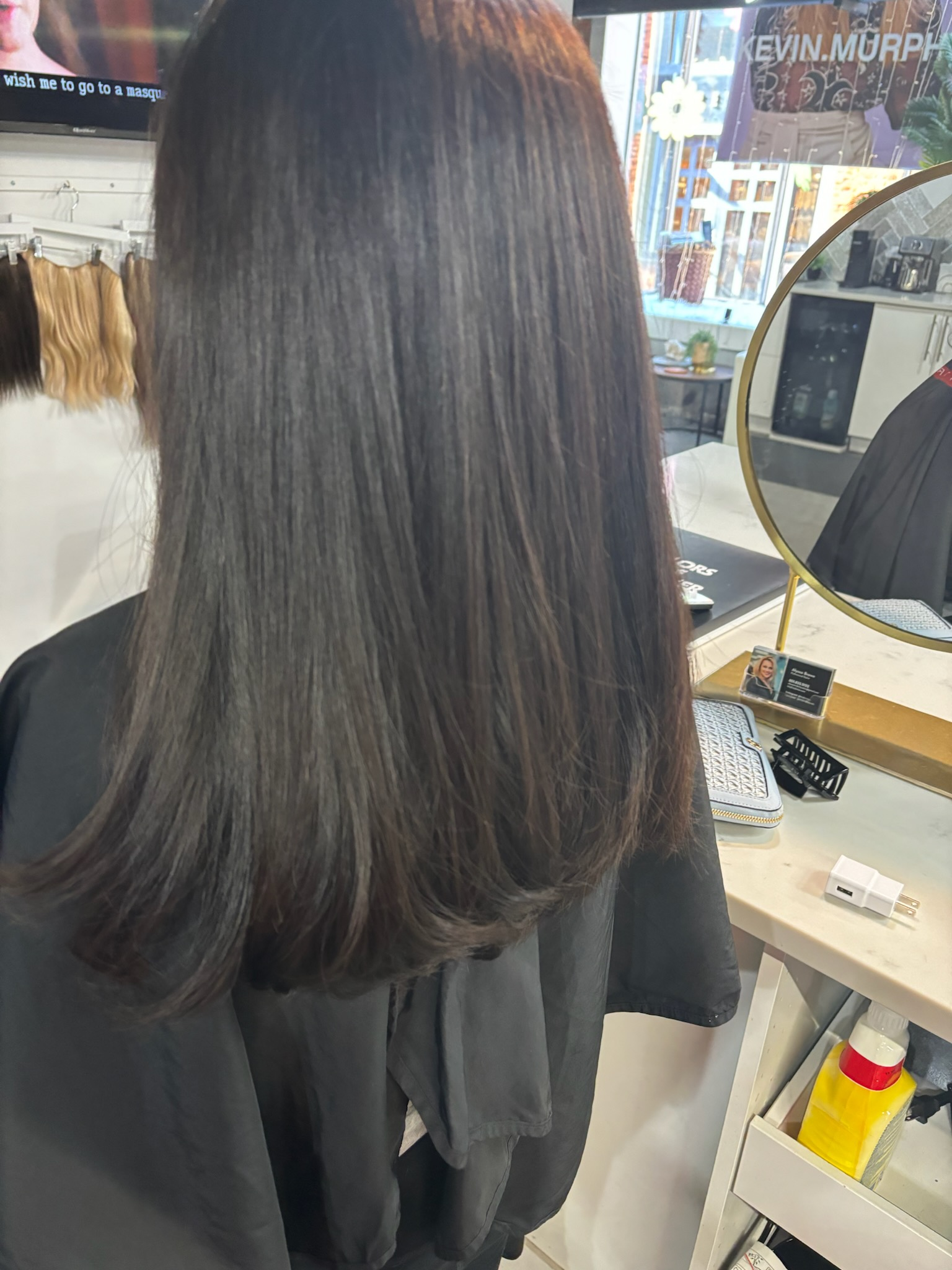 Long straight dark hair viewed from behind in a salon chair, with a mirror and styling tools nearby.