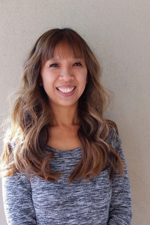 Smiling woman with long wavy hair wearing a gray top against a plain beige wall