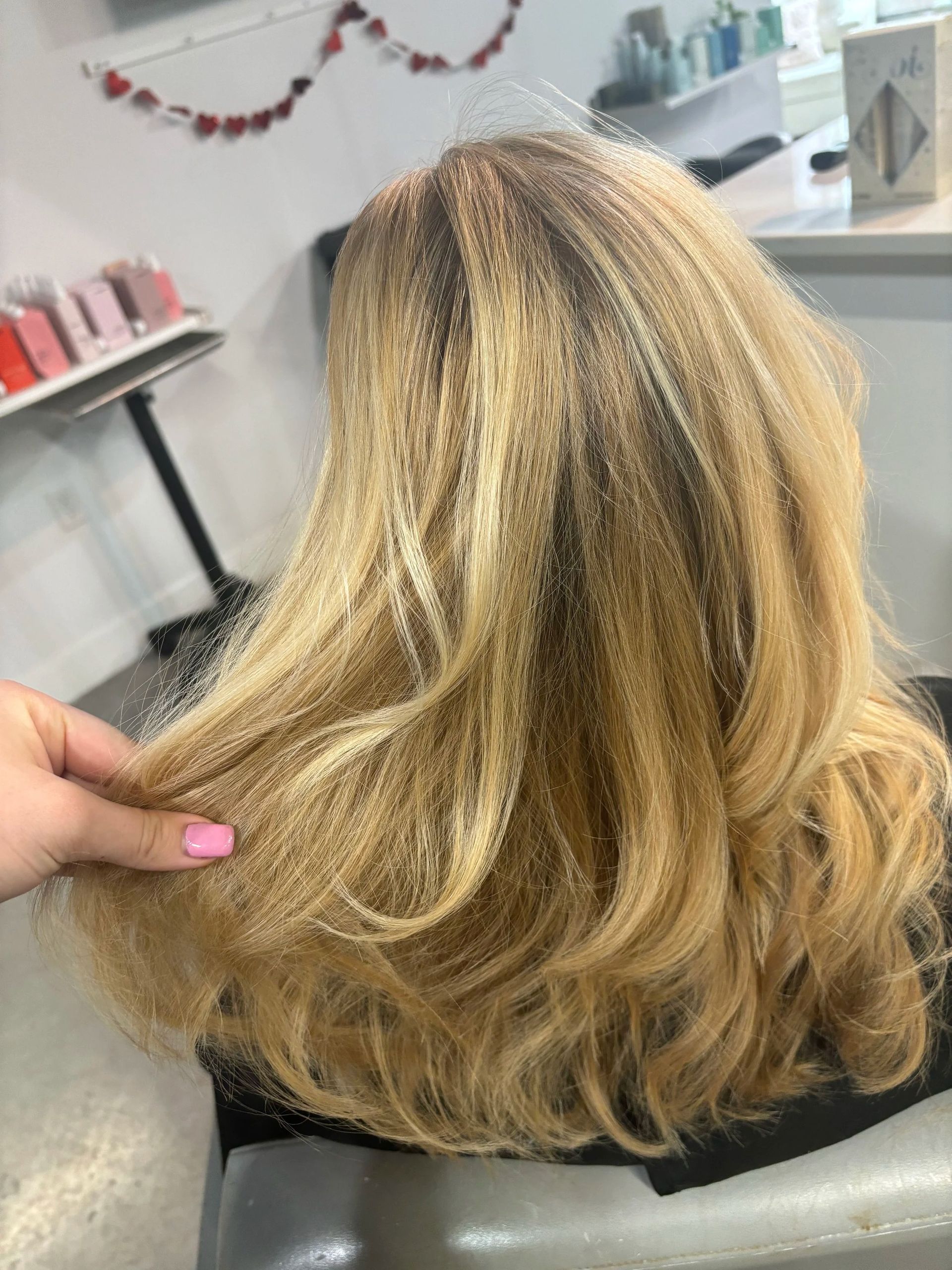 Blonde wavy shoulder-length hair being held up in a salon chair, showing subtle highlights.