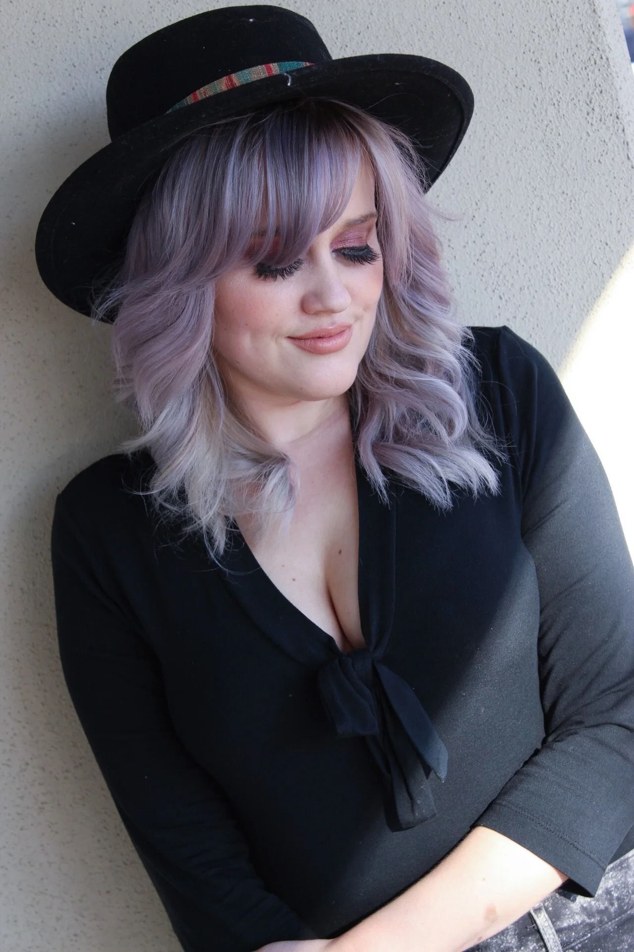 Person in a black hat and top with lilac hair, looking down against a light wall.
