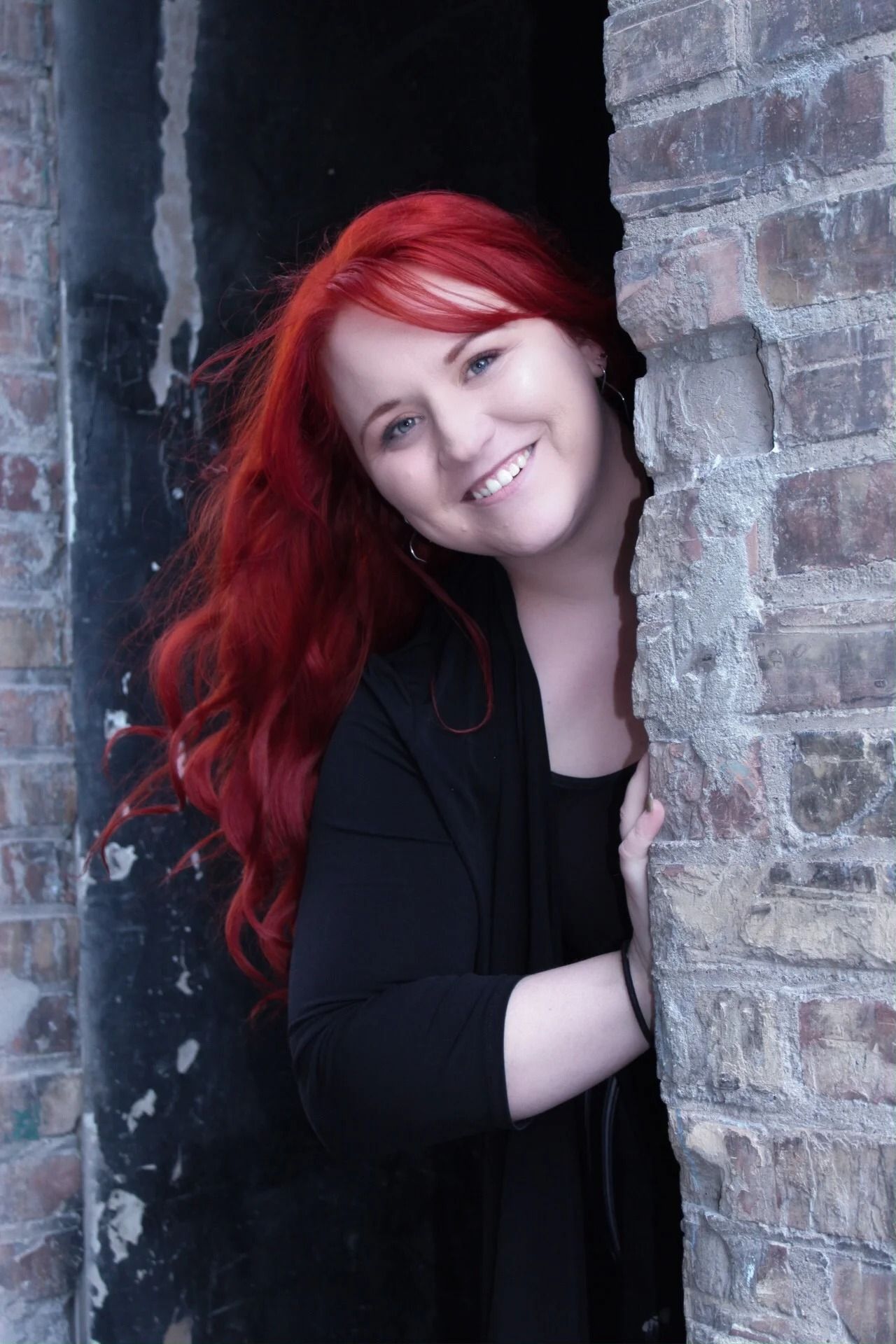 Smiling person with bright red hair peeks from behind a brick wall.