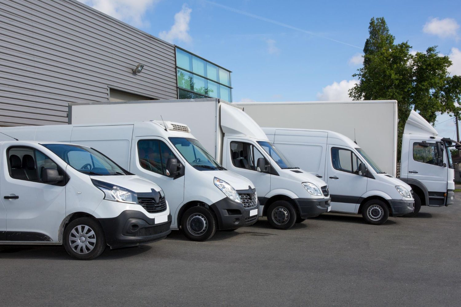 A row of white delivery vans parked in front of a modern industrial warehouse building under a blue sky.