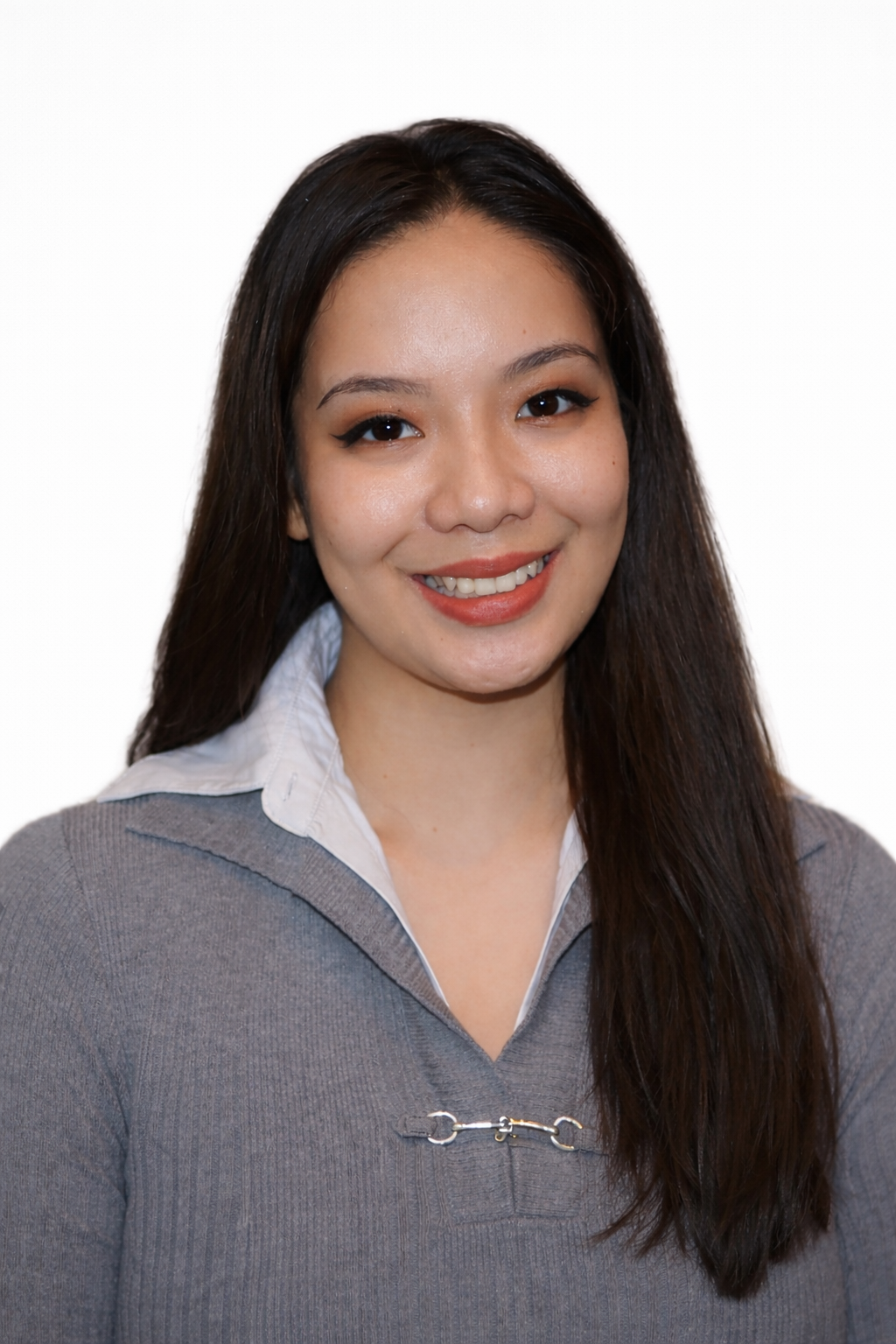 A person with long dark hair smiles while wearing a grey top with a white collar against a plain white background.