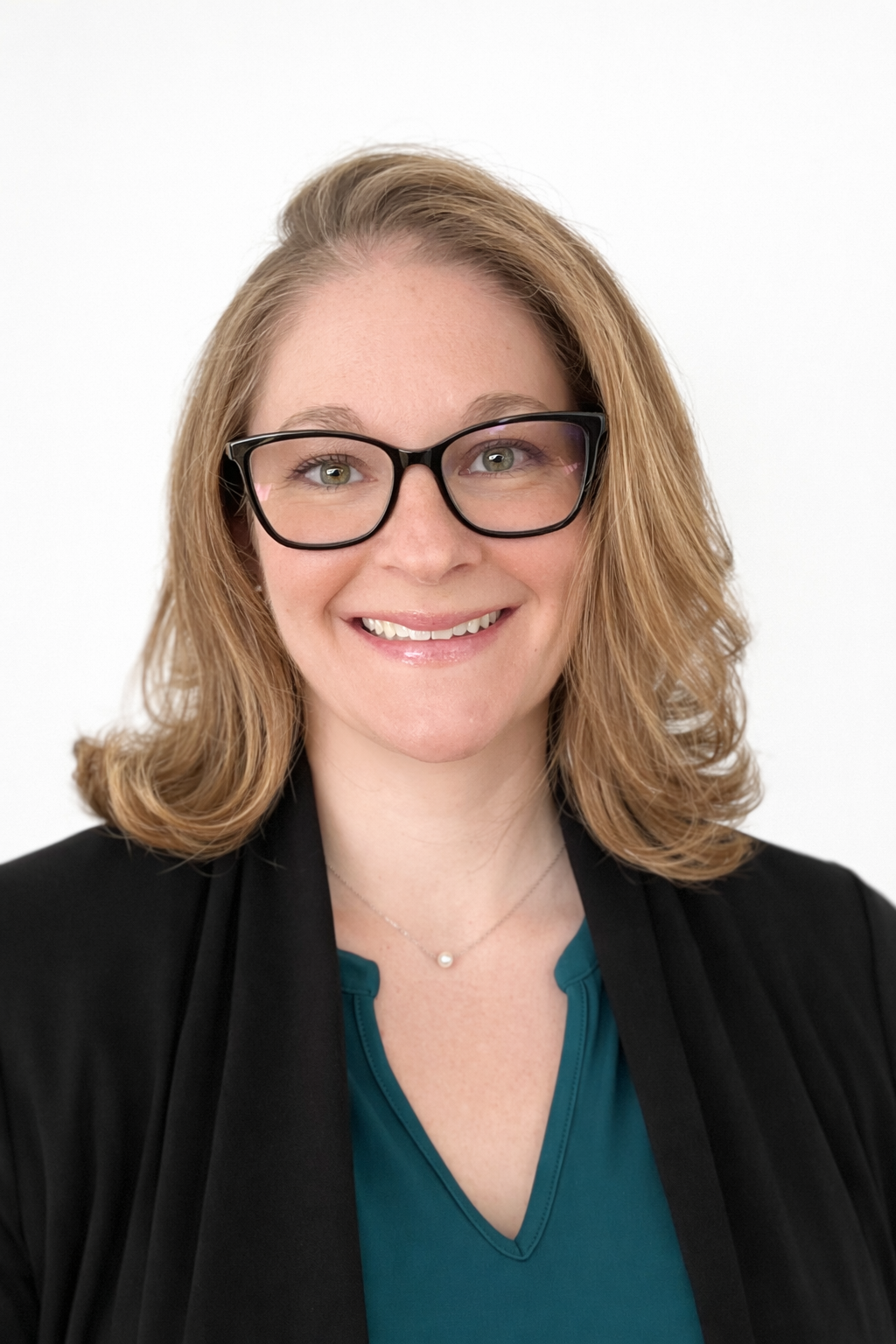 A person with shoulder-length hair and glasses smiles while wearing a teal top and a black open blazer against a white wall.