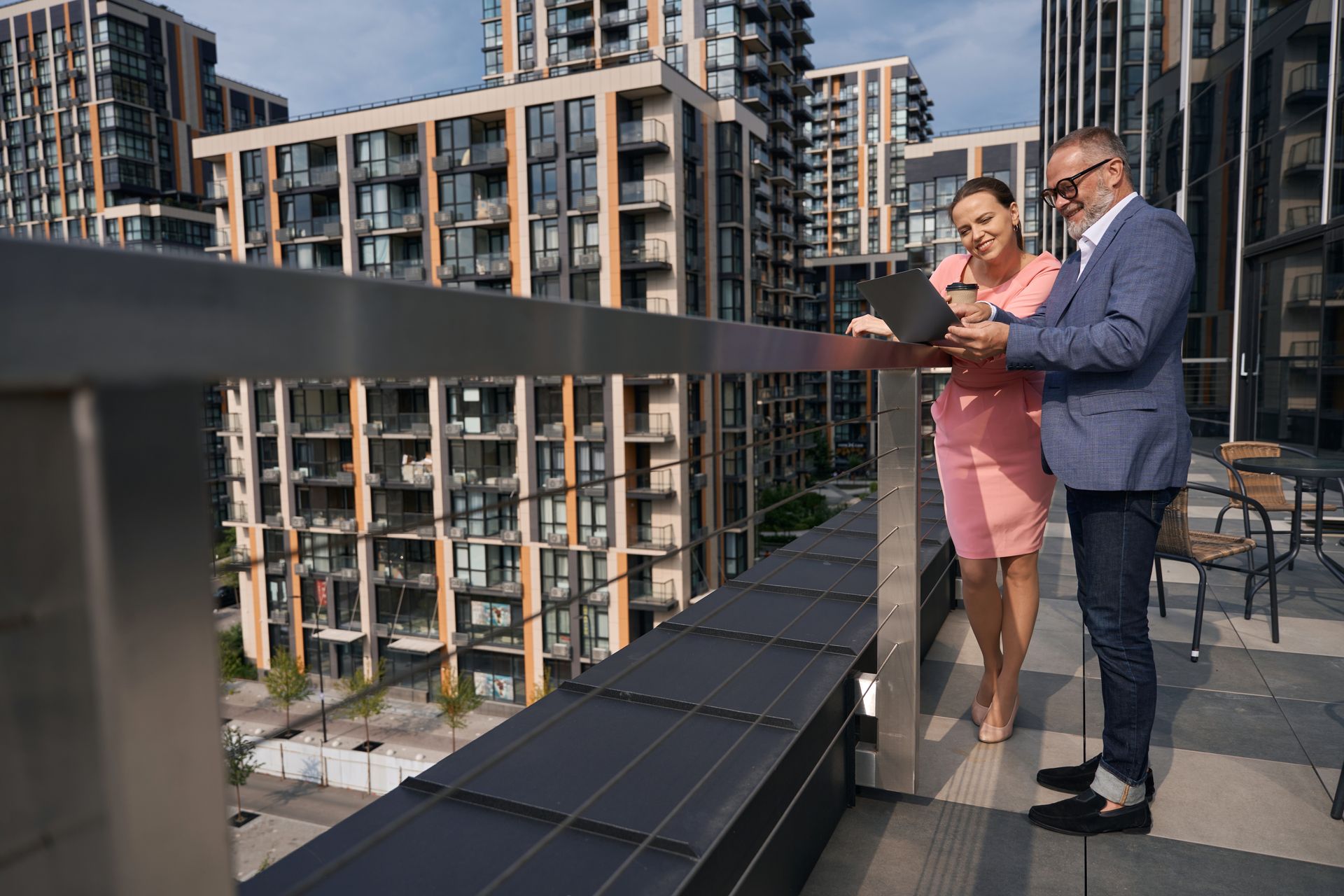 A professional man and woman look at a laptop together on a high-rise balcony overlooking city apartment buildings.