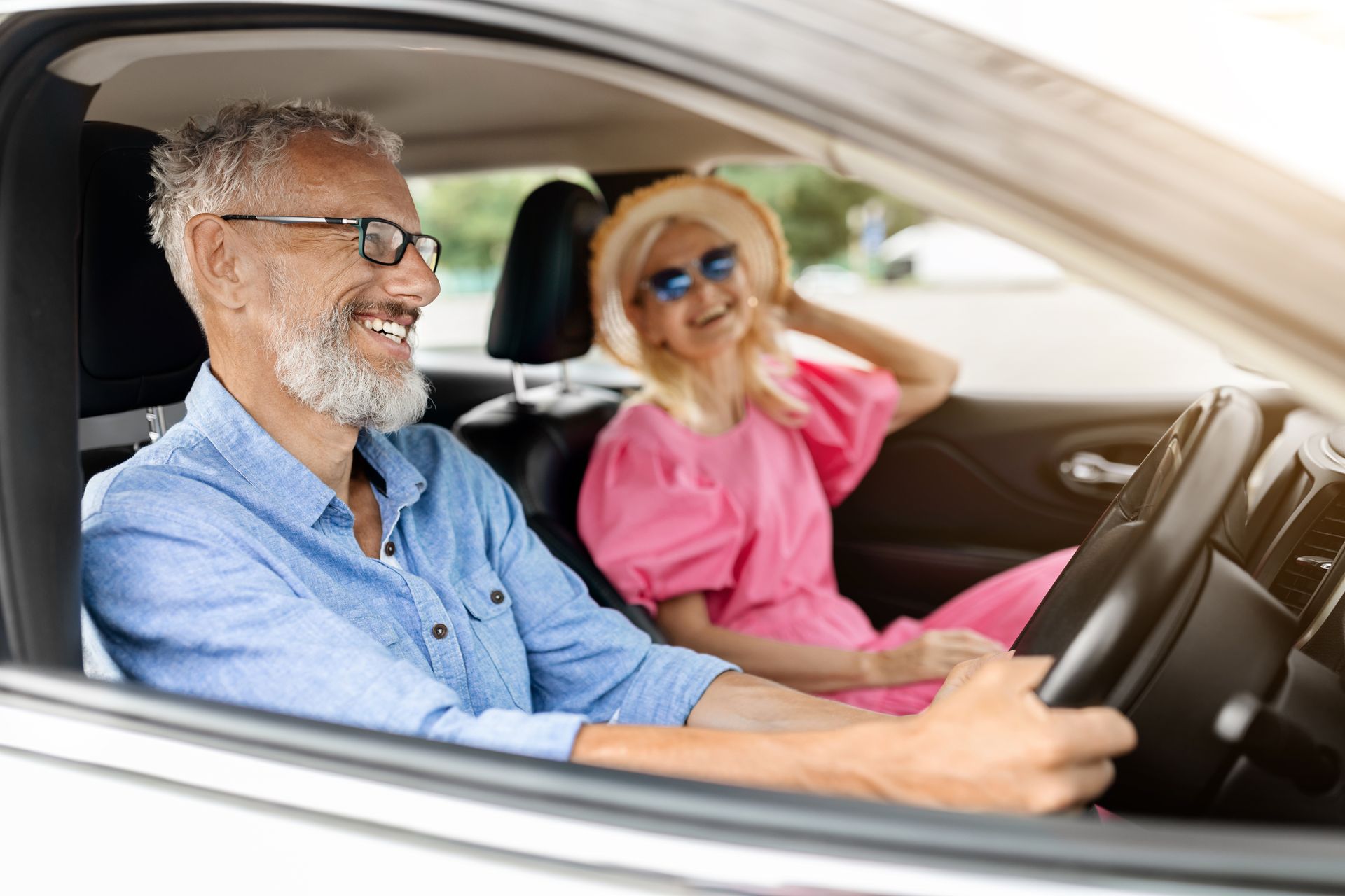 Two smiling people in a car; a driver wearing a blue shirt and glasses, and a passenger wearing a pink top and sun hat.