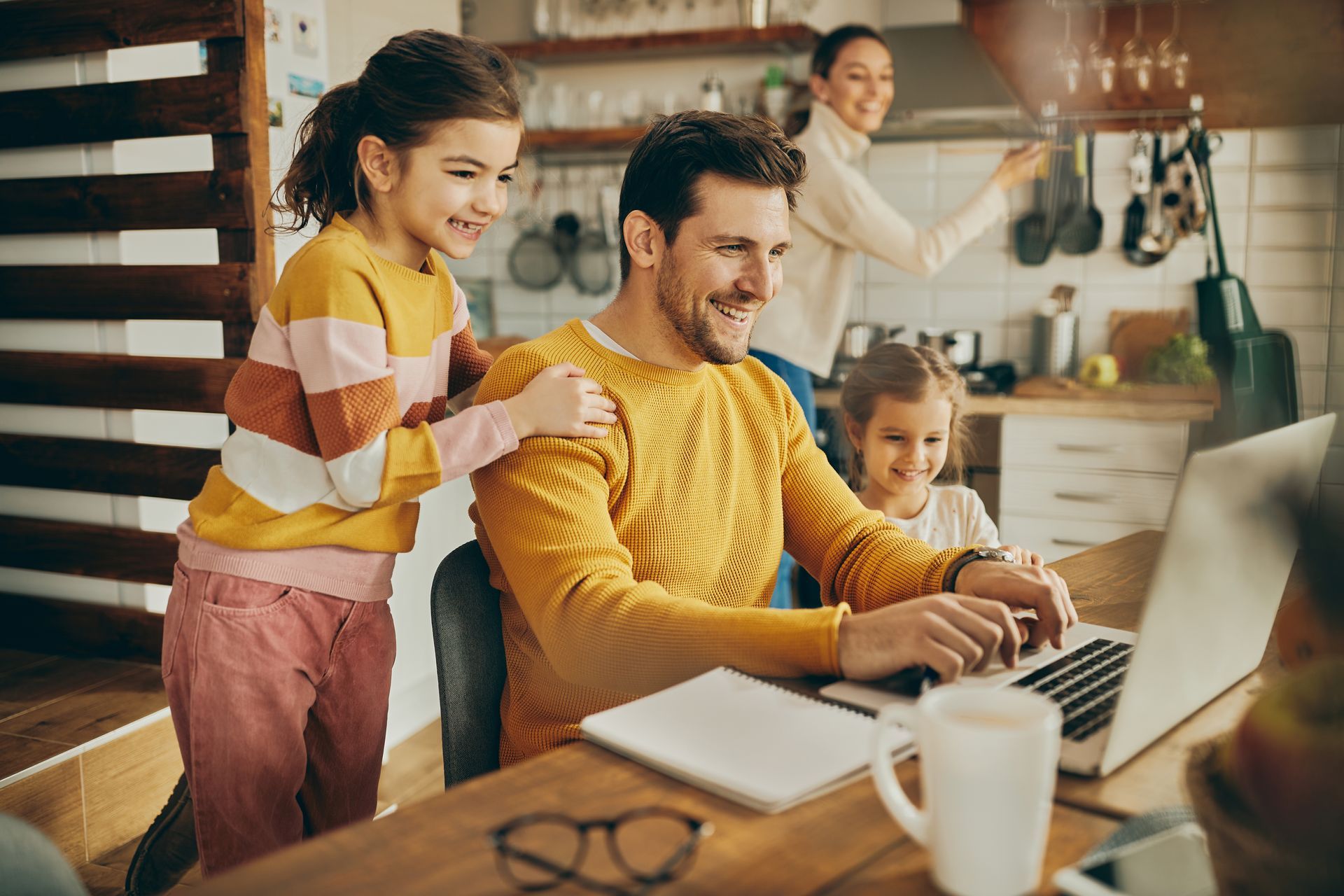 A person wearing a yellow sweater works on a laptop at a table with two children, while another person stands nearby.