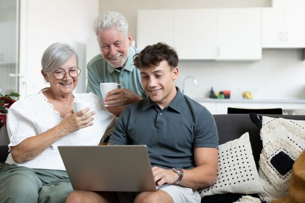 A young adult and two older adults sit on a couch in a bright room, looking together at a laptop screen.