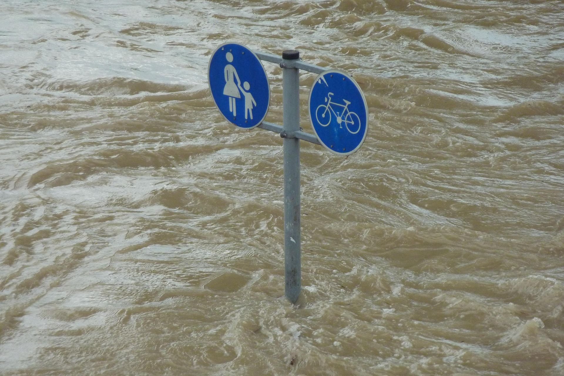 A blue pedestrian and bicycle path sign stands partially submerged in rushing, muddy floodwaters.