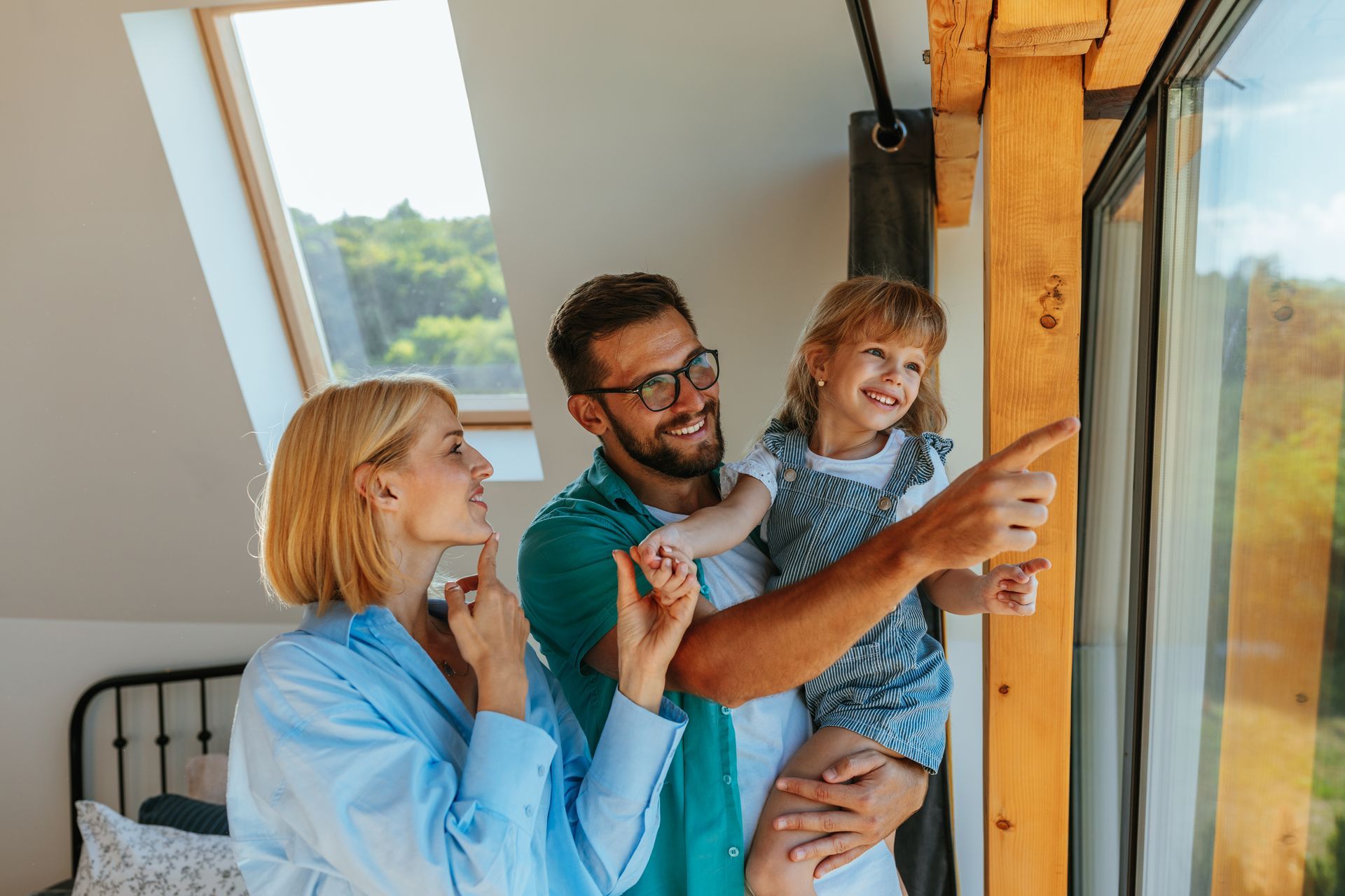 A family stands by a bright window, pointing outside with smiling expressions in a sunlit room.