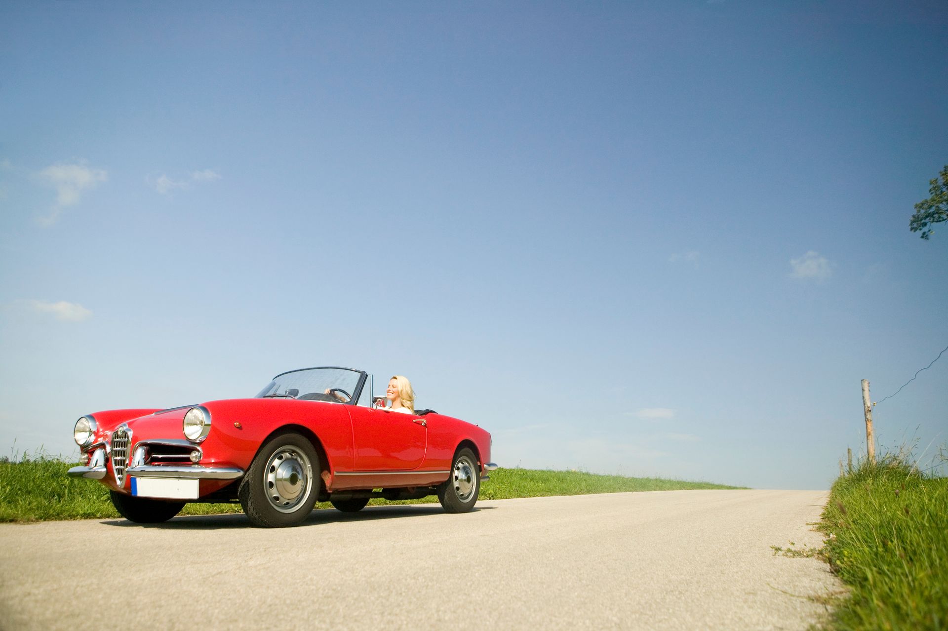 A bright red vintage Alfa Romeo convertible drives along a narrow, paved country road under a clear blue sky.