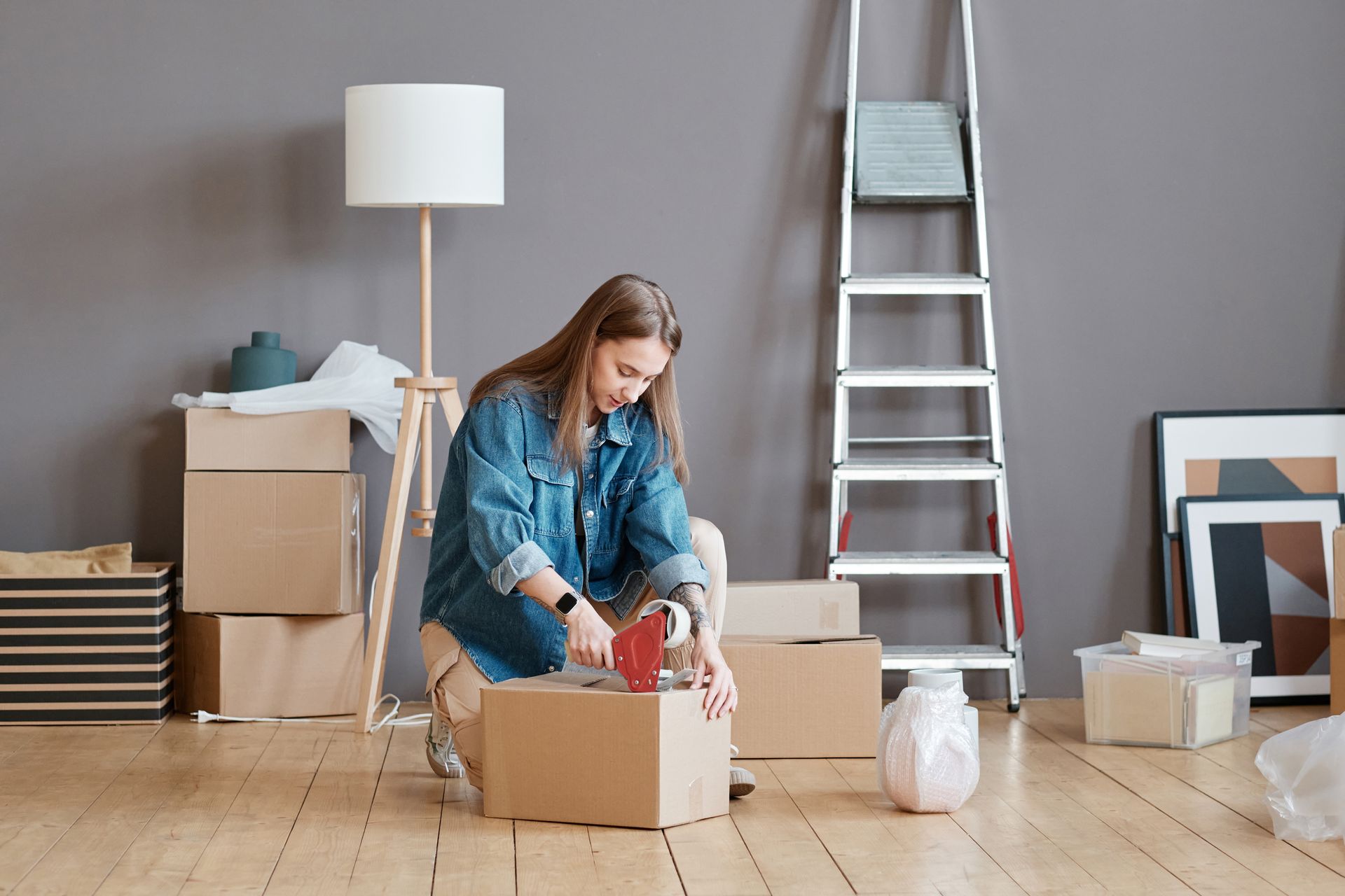 A person kneeling on a wooden floor, sealing a cardboard moving box with packing tape in a room with boxes and a ladder.